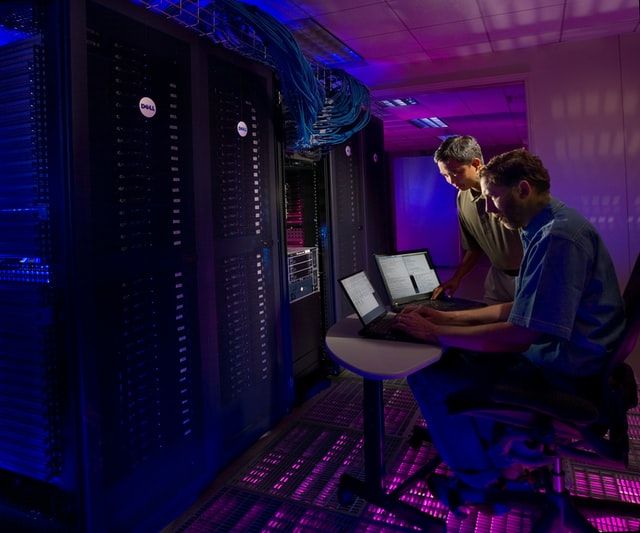 Two men in a data centre working on laptops beside several racks of servers