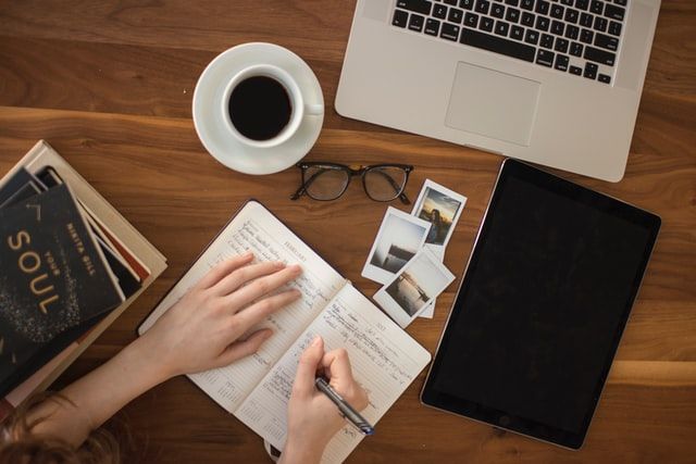 An author writing in a notebook, with coffee and a laptop also on the desk